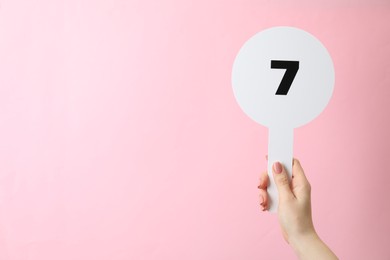 Woman holding auction paddle with number 7 on pink background, closeup. Space for text