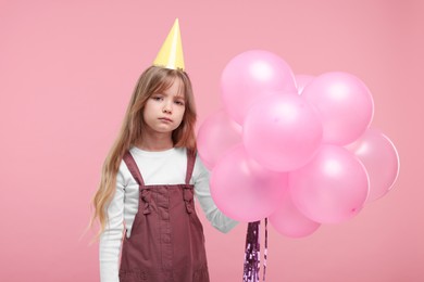 Photo of Bored little girl in party hat with bunch of balloons on pink background
