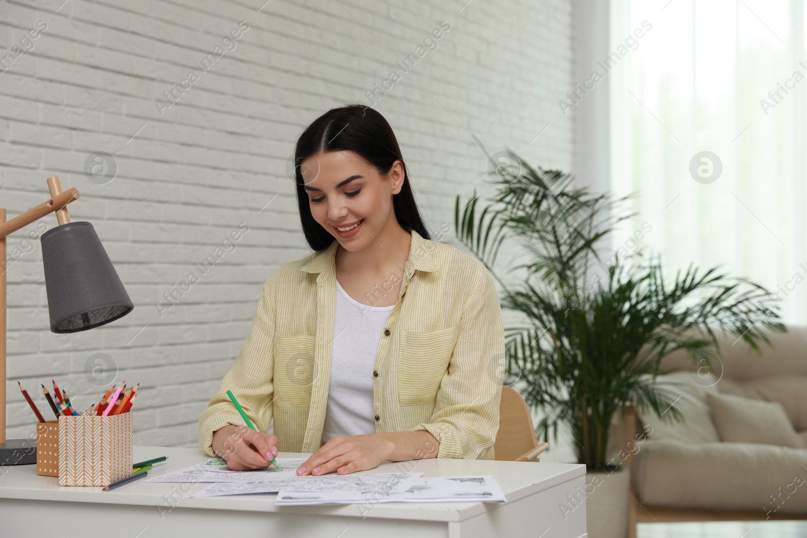 Photo of Young woman coloring antistress page at desk indoors