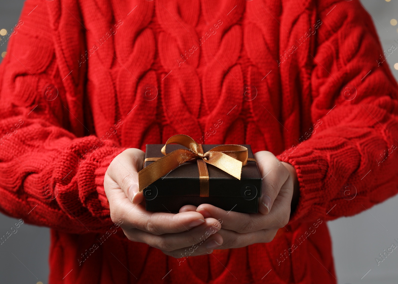 Photo of Christmas present. Woman holding gift box against grey background with blurred lights, closeup