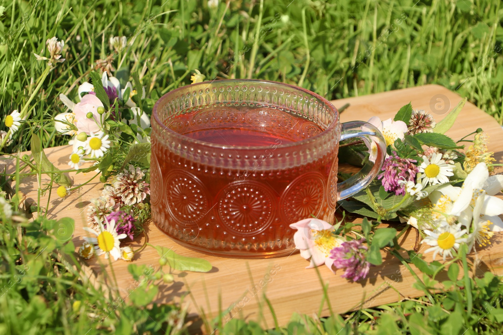 Photo of Ornate glass cup of tea, different wildflowers and herbs on wooden board in meadow