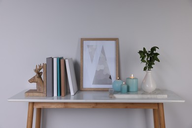Photo of Burning candles, books, picture and vase with green branches on console table near white wall