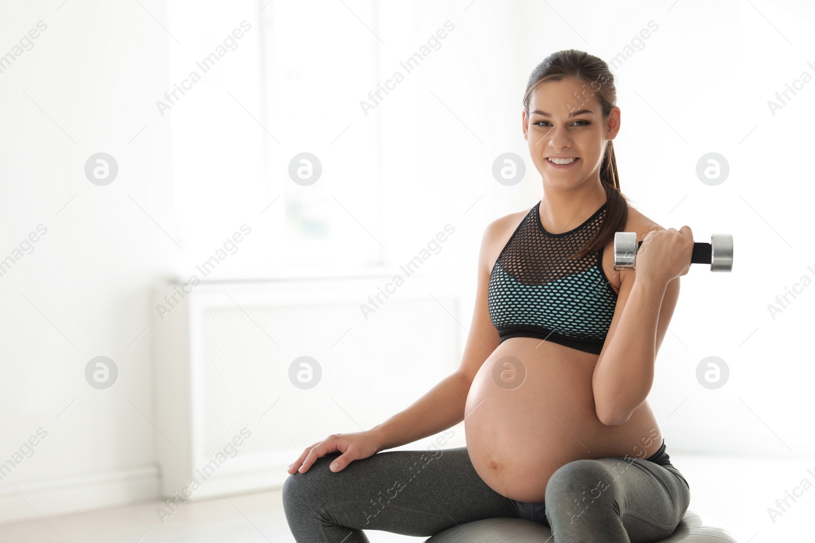 Photo of Young pregnant woman in fitness clothes lifting dumbbell at home. Space for text