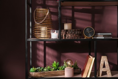 Photo of Stylish wicker candle holder, night lamp with incandescent bulb and decor on shelving unit near brown wall