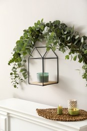 Beautiful garland made of eucalyptus branches and aromatic candle hanging above mantelpiece indoors