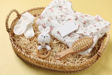 Different baby accessories, clothes and blank card in wicker basket on yellow background, closeup