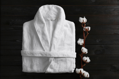 Photo of Clean folded bathrobe and branch of cotton on black wooden background, flat lay