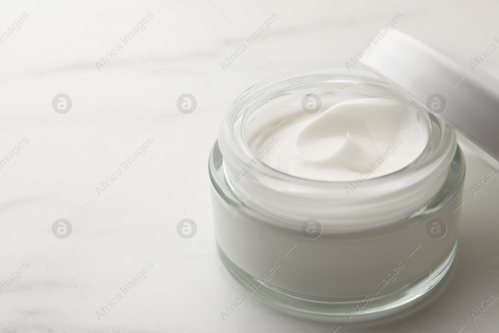 Photo of Glass jar of face cream on white marble table, closeup. Space for text