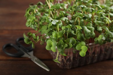 Photo of Fresh radish microgreens in plastic container and scissors on wooden table, closeup