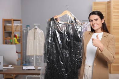 Photo of Dry-cleaning service. Happy woman holding hanger with dress in plastic bag and showing thumb up indoors, space for text