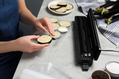 Photo of Woman packing cut eggplant into plastic bag using vacuum sealer on light grey marble table, closeup