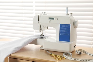 Photo of White sewing machine, scissors, measuring tape and fabric on wooden table indoors