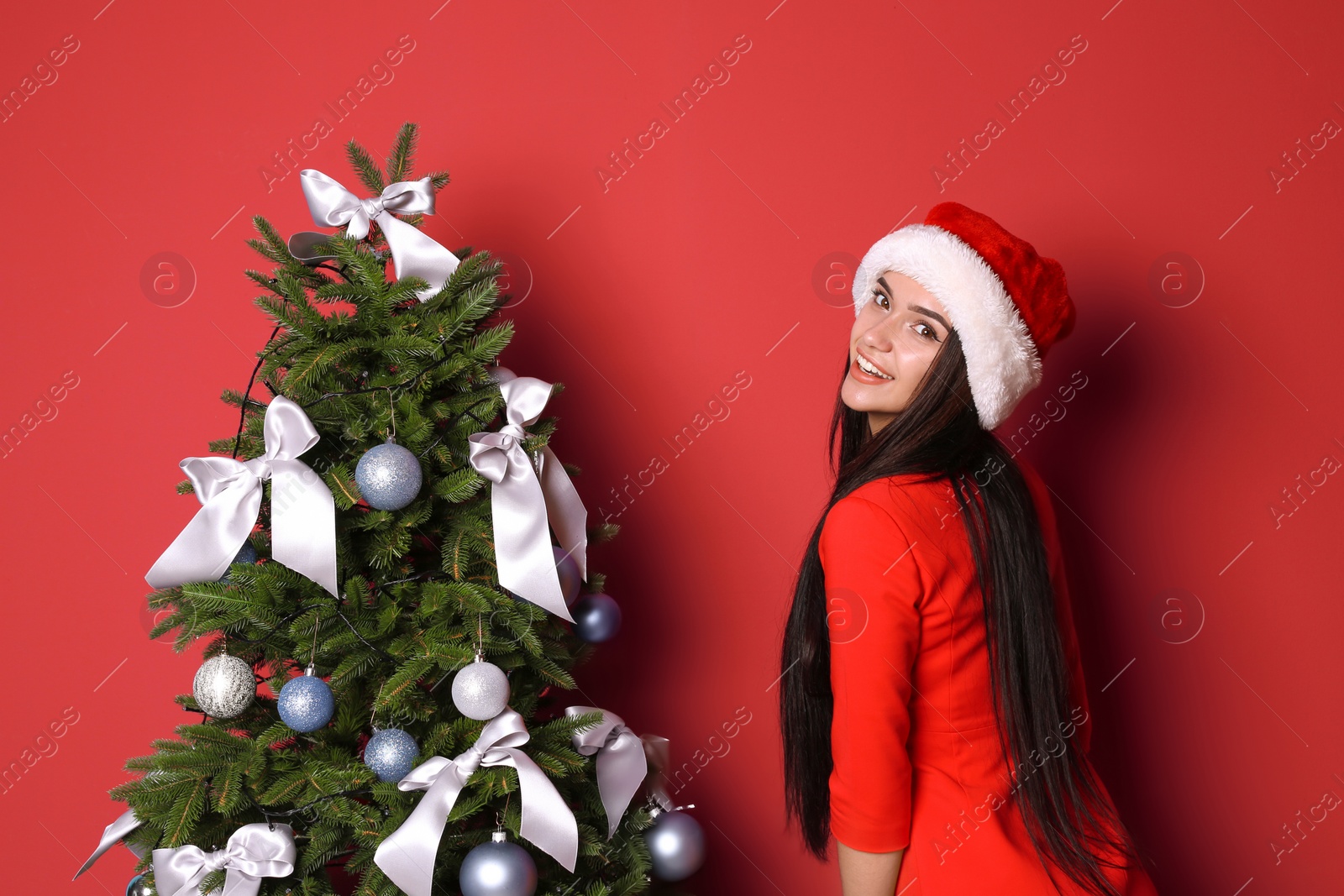 Photo of Beautiful young woman in Santa hat near Christmas tree on color background