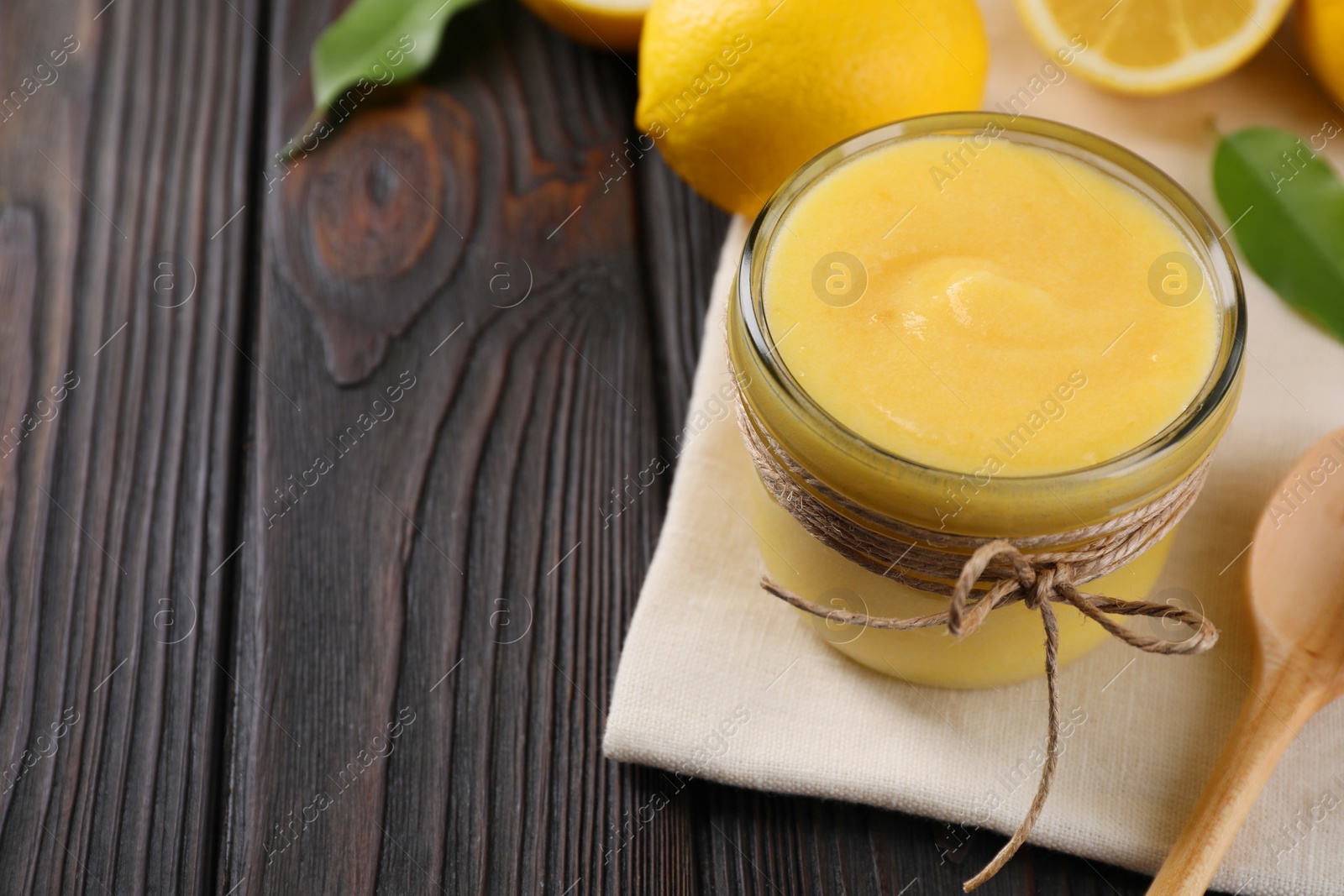 Photo of Delicious lemon curd in glass jar, fresh citrus fruit and spoon on wooden table, space for text