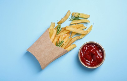 Photo of Paper cup with French fries, rosemary and ketchup on light blue table, flat lay