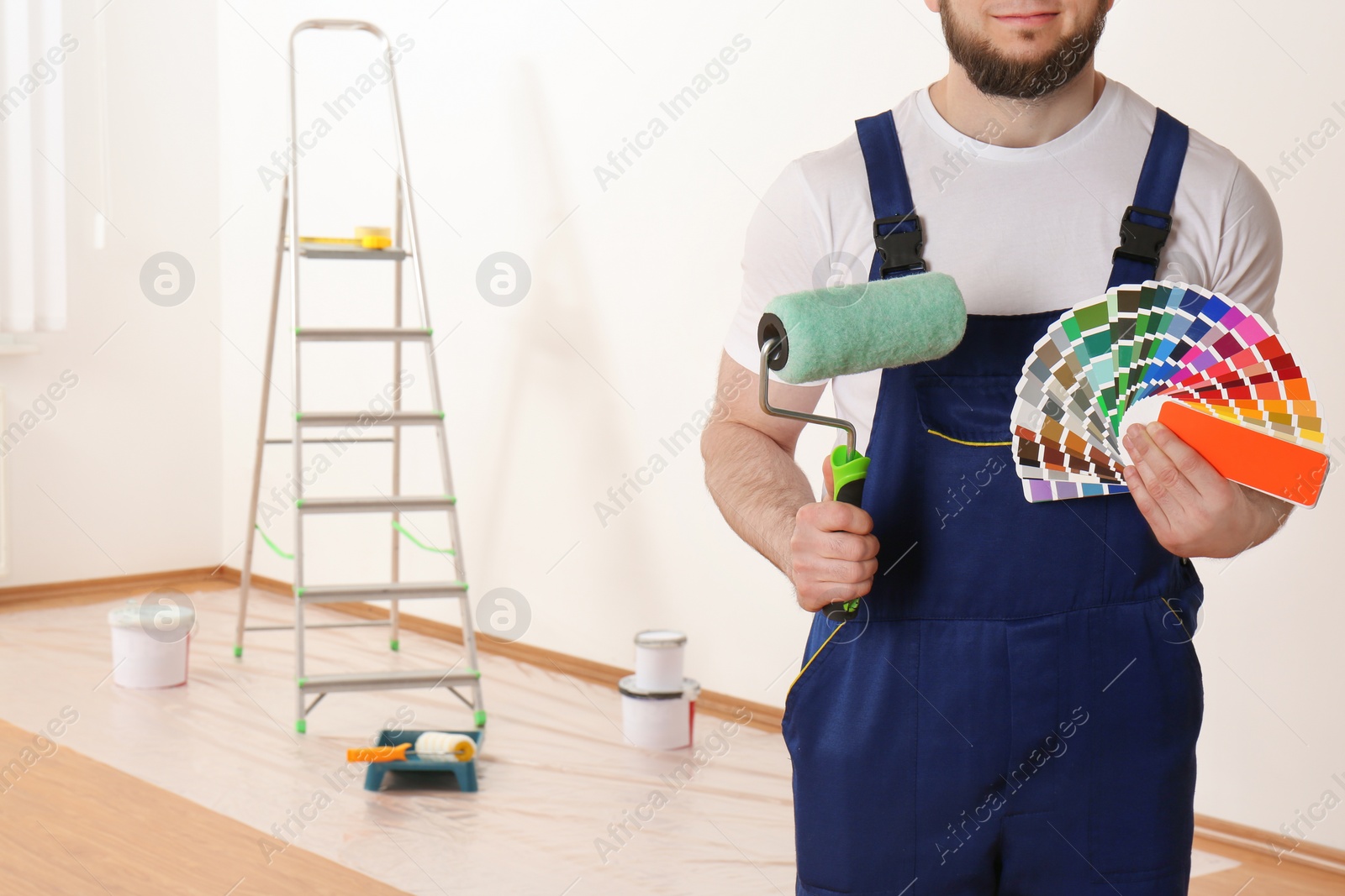 Photo of Male decorator with paint roller and color palette samples in empty room, closeup. Space for text