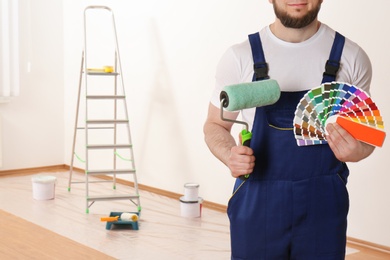 Photo of Male decorator with paint roller and color palette samples in empty room, closeup. Space for text