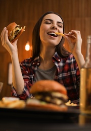 Young woman eating French fries and tasty burger in cafe