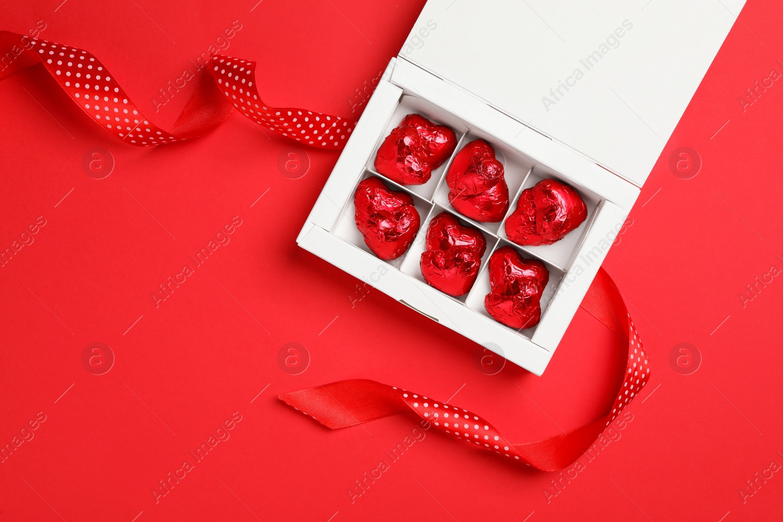 Photo of Tasty chocolate heart shaped candies in white box with ribbon on red background, flat lay