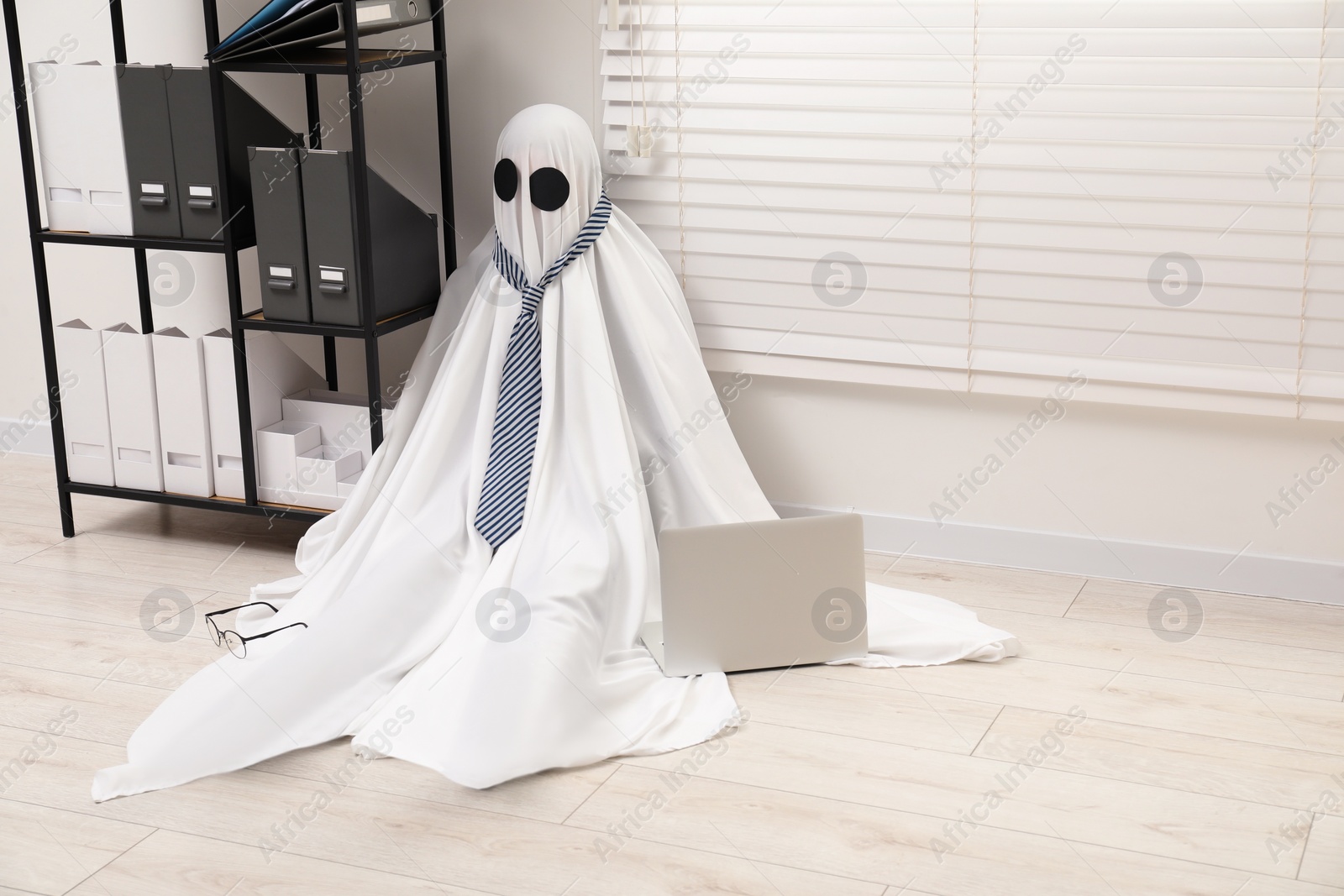 Photo of Overworked ghost. Man in white sheet with laptop on floor in office, space for text