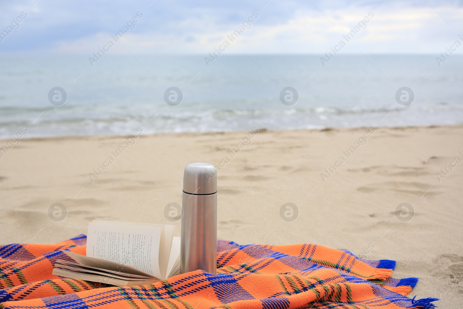 Photo of Metallic thermos with hot drink, open book and plaid on sandy beach near sea, space for text