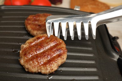 Photo of Taking delicious hamburger patty with tongs from grill pan, closeup