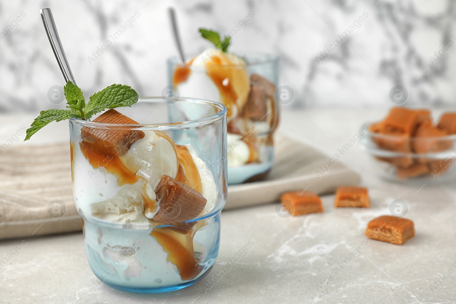 Photo of Glass of delicious ice cream with caramel sauce, candies and mint on light grey marble table. Space for text