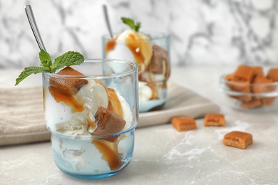 Glass of delicious ice cream with caramel sauce, candies and mint on light grey marble table. Space for text