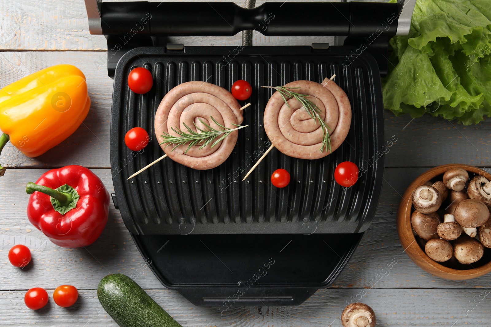 Photo of Electric grill with homemade sausages, rosemary and vegetables on rustic wooden table, flat lay