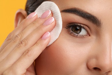 Beautiful woman removing makeup with cotton pad on orange background, closeup