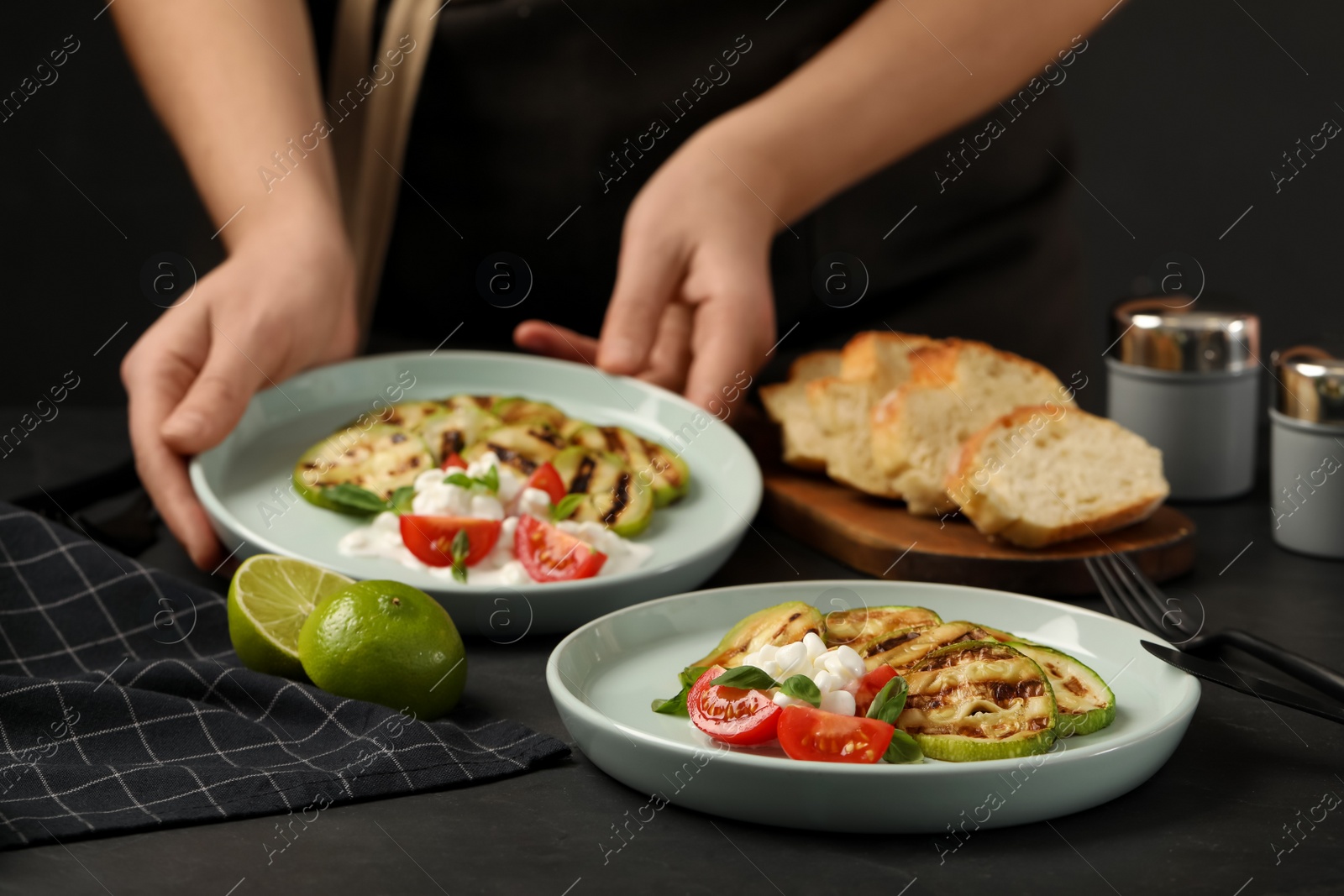 Photo of Woman serving delicious grilled zucchini slices with cottage cheese and tomatoes at black table