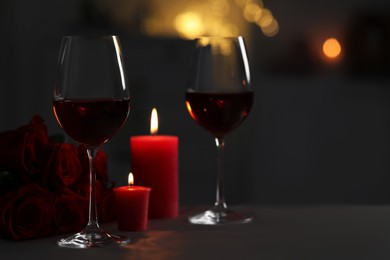 Photo of Glasses of red wine, burning candles and rose flowers on grey table against blurred lights, space for text. Romantic atmosphere