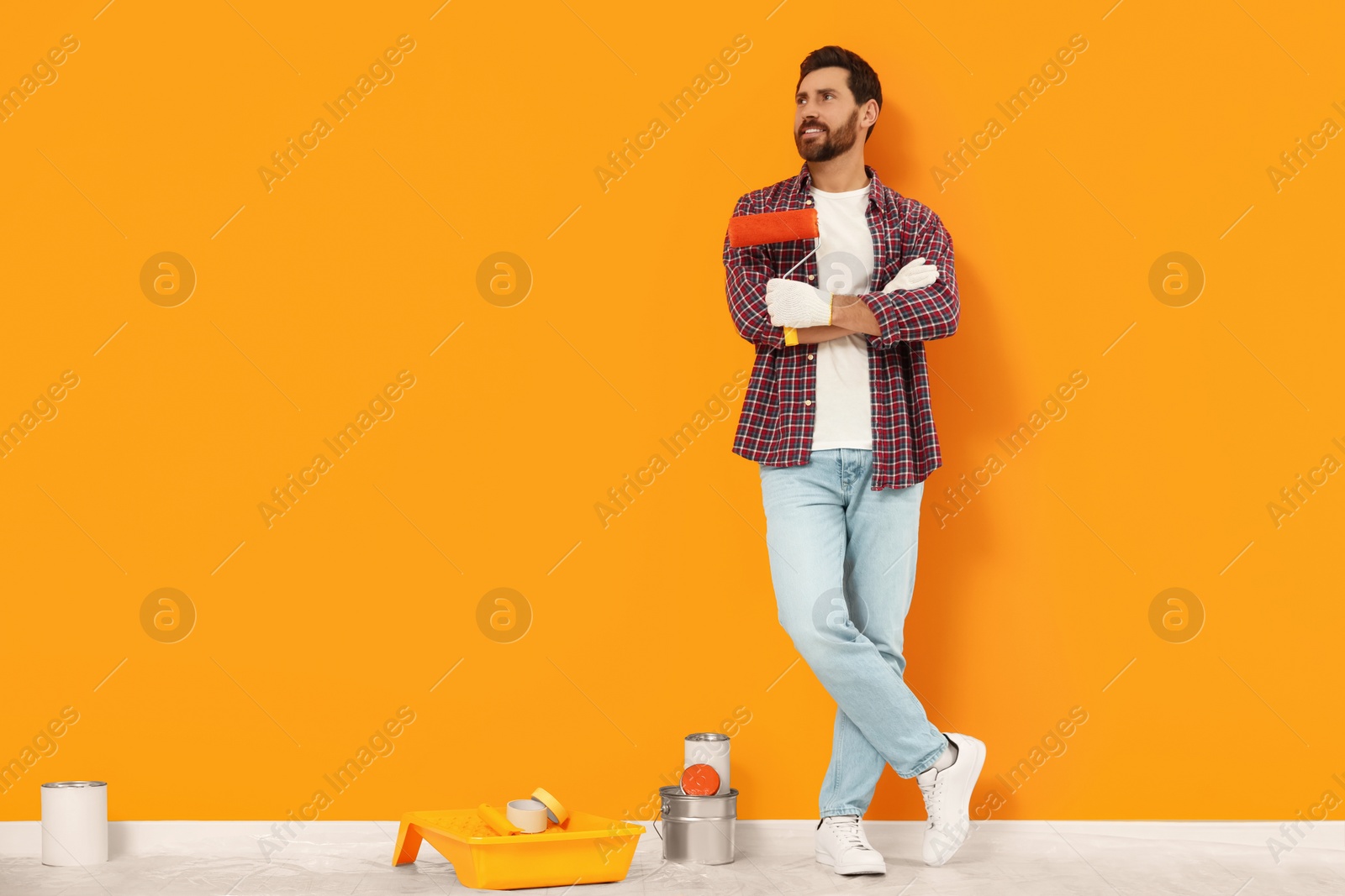 Photo of Designer with roller and painting equipment on floor near freshly painted orange wall indoors, space for text