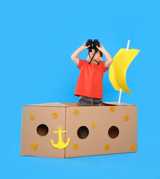 Cute little child playing with binoculars and cardboard ship on light blue background