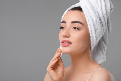 Beautiful woman in terry towel removing makeup with cotton pad on gray background, space for text