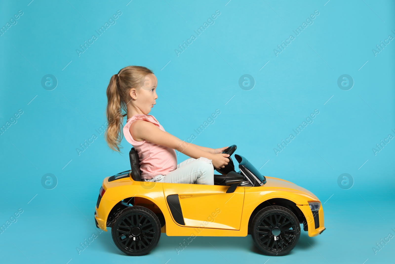Photo of Cute little girl driving children's electric toy car on light blue background
