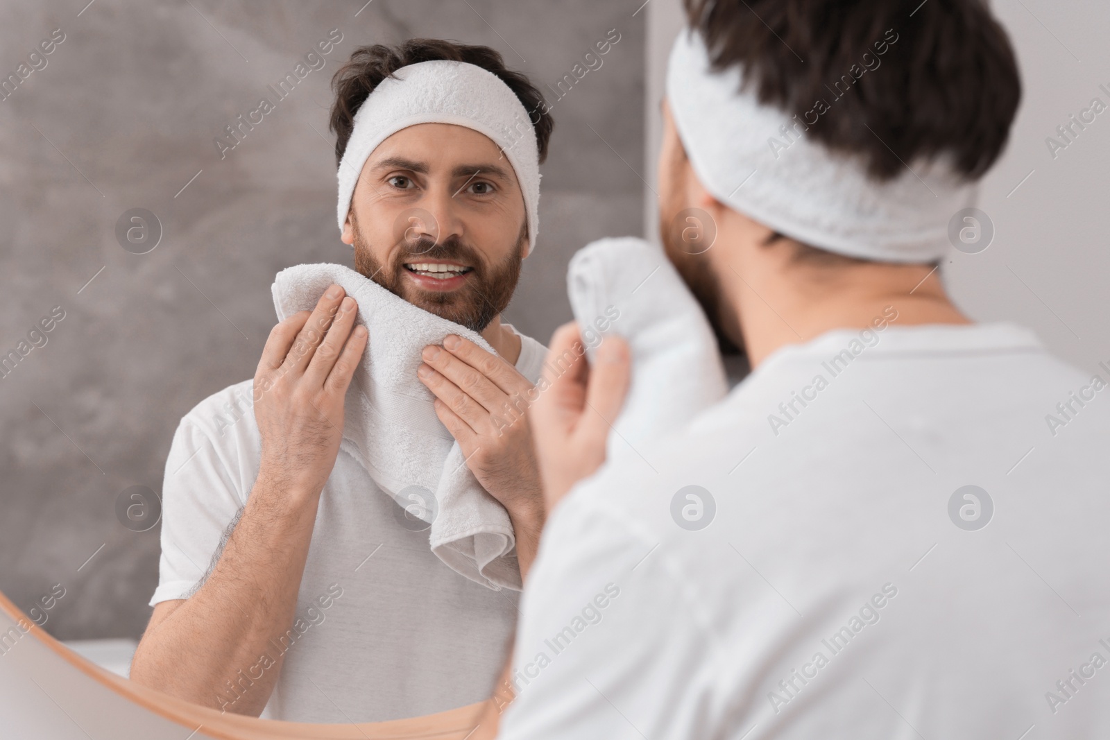 Photo of Washing face. Man with headband and towel near mirror in bathroom