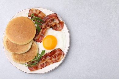 Plate with tasty pancakes, fried egg, arugula and bacon on light grey textured table, top view. Space for text