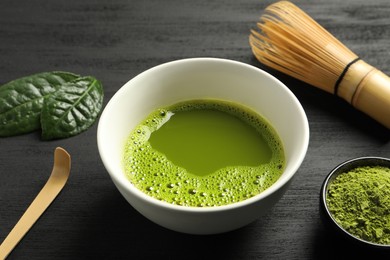 Cup of fresh matcha tea, green powder and bamboo whisk on black wooden table, closeup