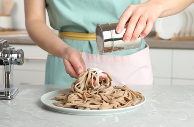 Woman sprinkling raw soba (buckwheat noodles) with flour at grey table in kitchen, closeup