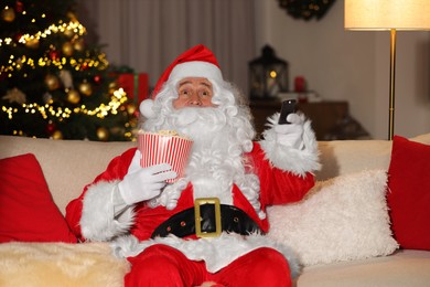 Photo of Merry Christmas. Santa Claus with popcorn bucket changing TV channels on sofa at home
