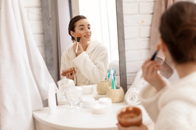 Young woman looking in mirror and applying natural scrub on face in bathroom