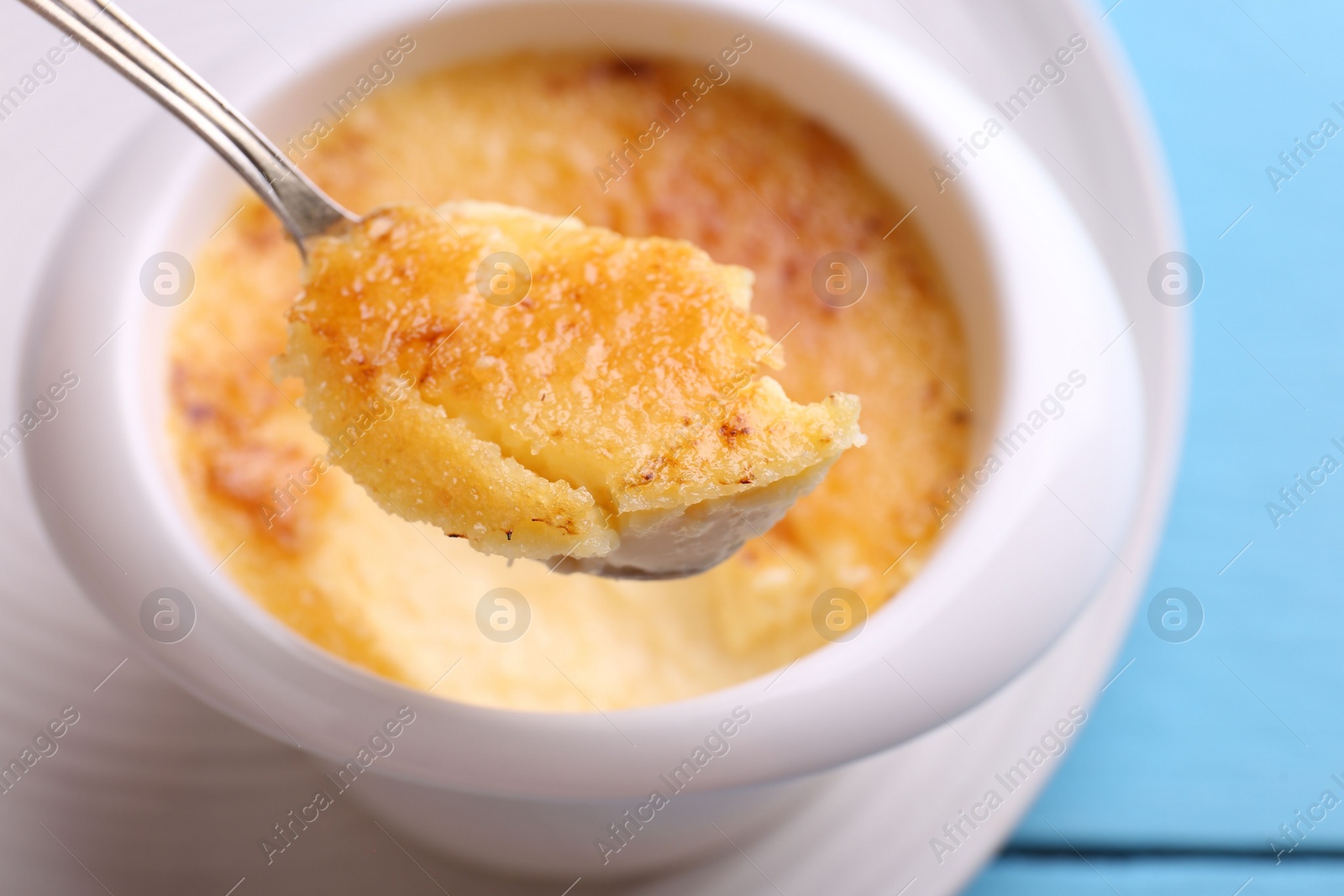 Photo of Taking delicious creme brulee with spoon from bowl at light blue table, closeup