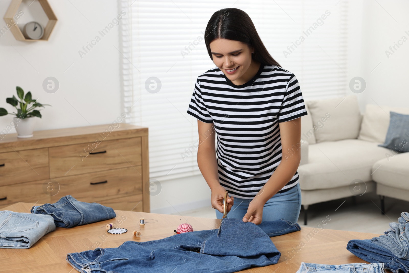 Photo of Young woman shortening jeans with scissors at wooden table indoors, space for text