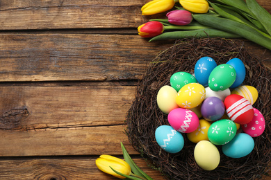 Photo of Colorful Easter eggs in decorative nest and tulips on wooden background, flat lay. Space for text