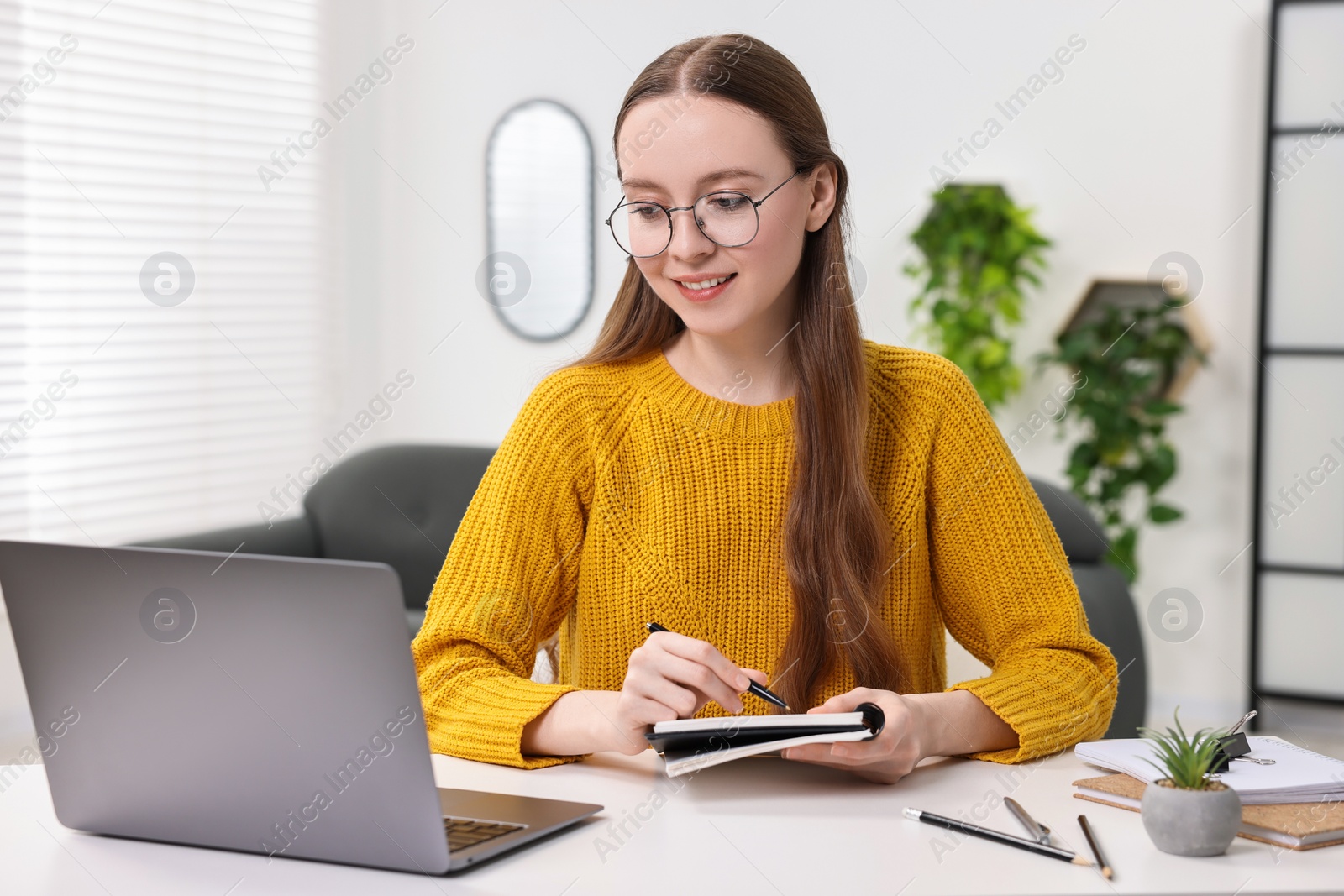 Photo of E-learning. Young woman taking notes during online lesson at white table indoors