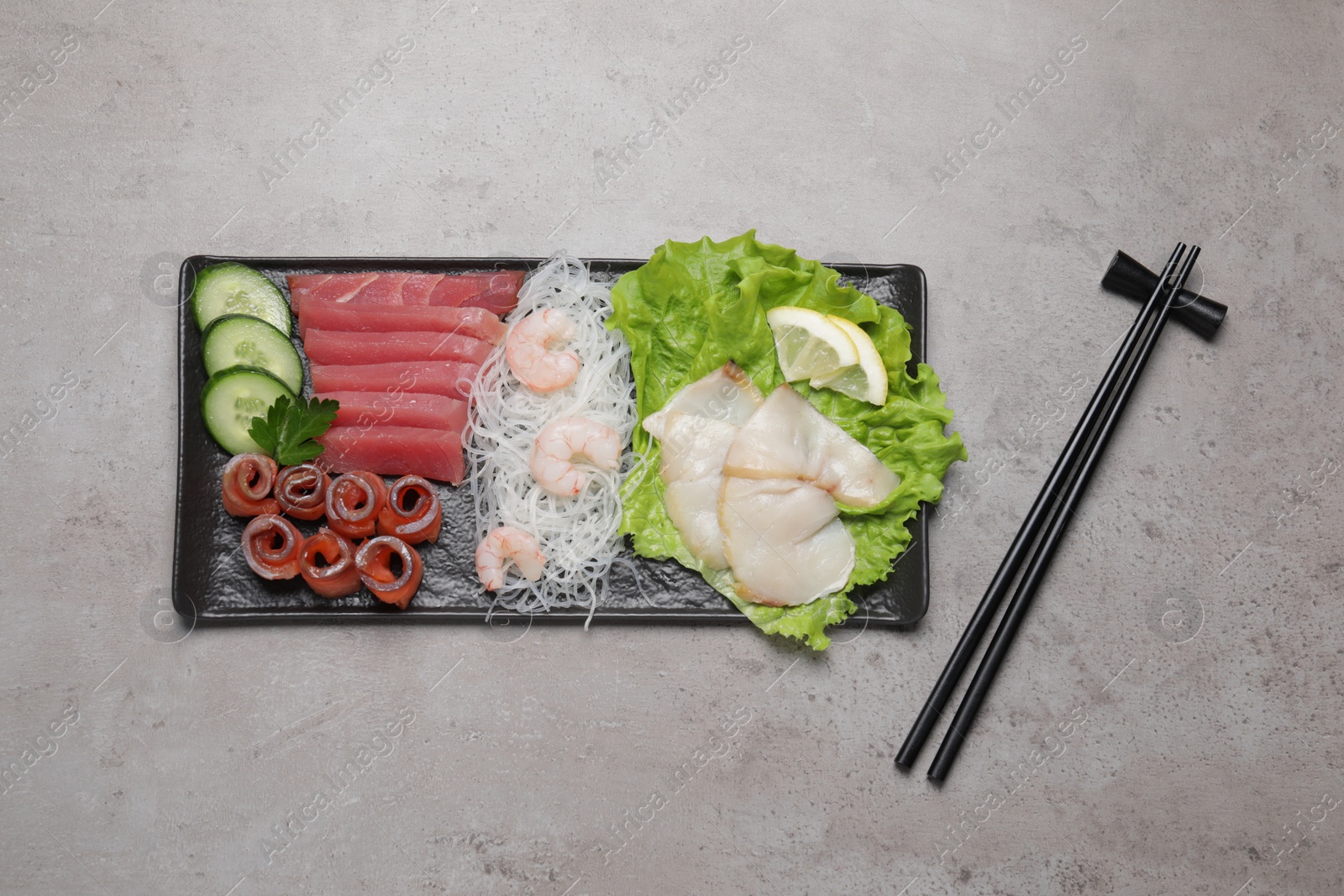 Photo of Sashimi set (raw slices of tuna, salmon, oily fish and shrimps) served with cucumber, lettuce, funchosa and lemon on light grey table, flat lay