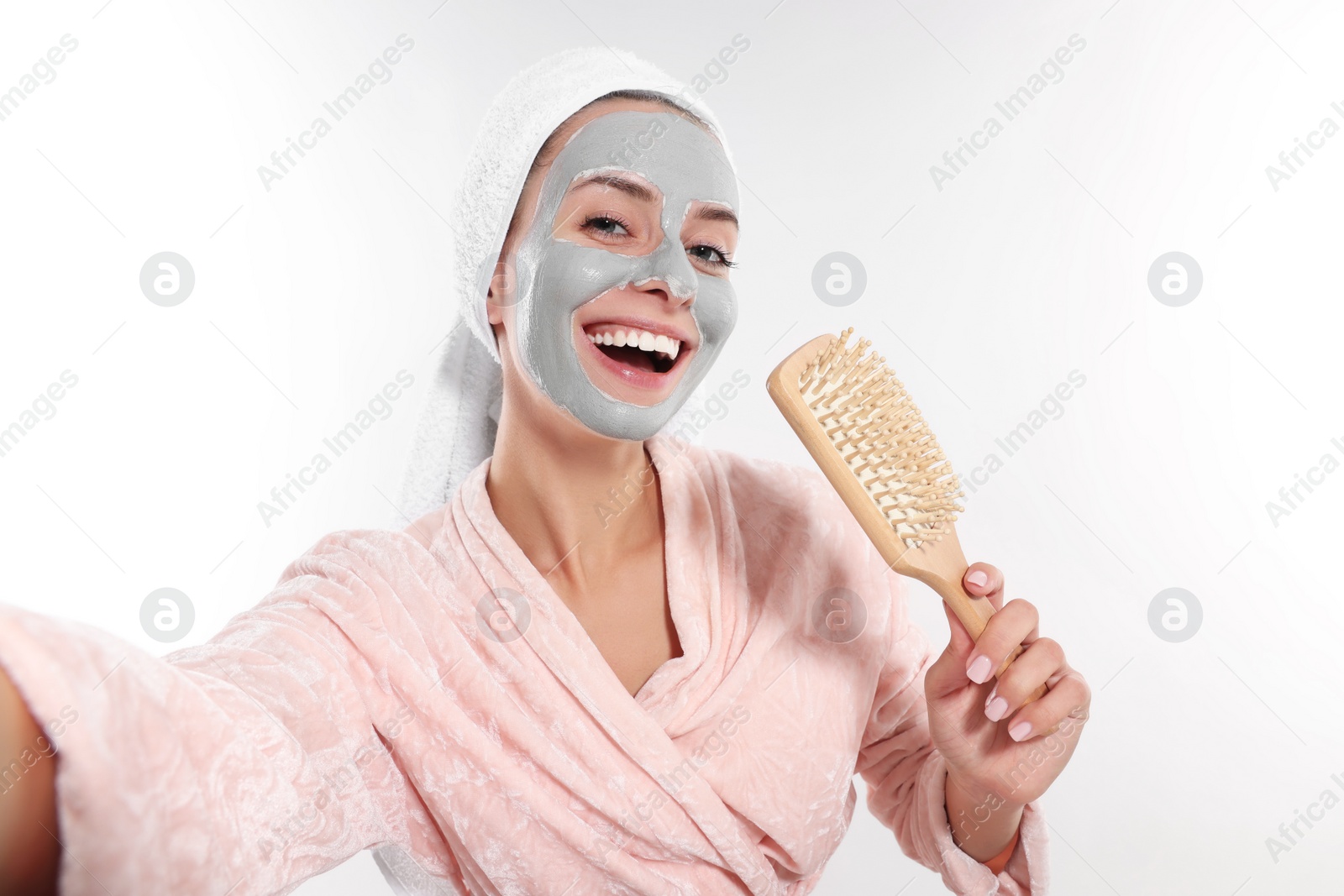 Photo of Woman with face mask and brush singing while taking selfie on white background. Spa treatments