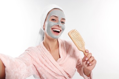 Woman with face mask and brush singing while taking selfie on white background. Spa treatments