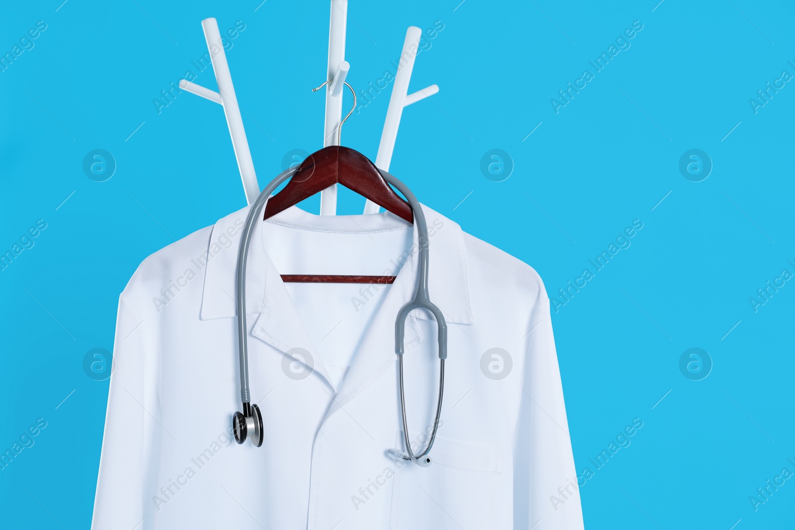 Photo of White medical uniform and stethoscope hanging on rack against light blue background. Space for text